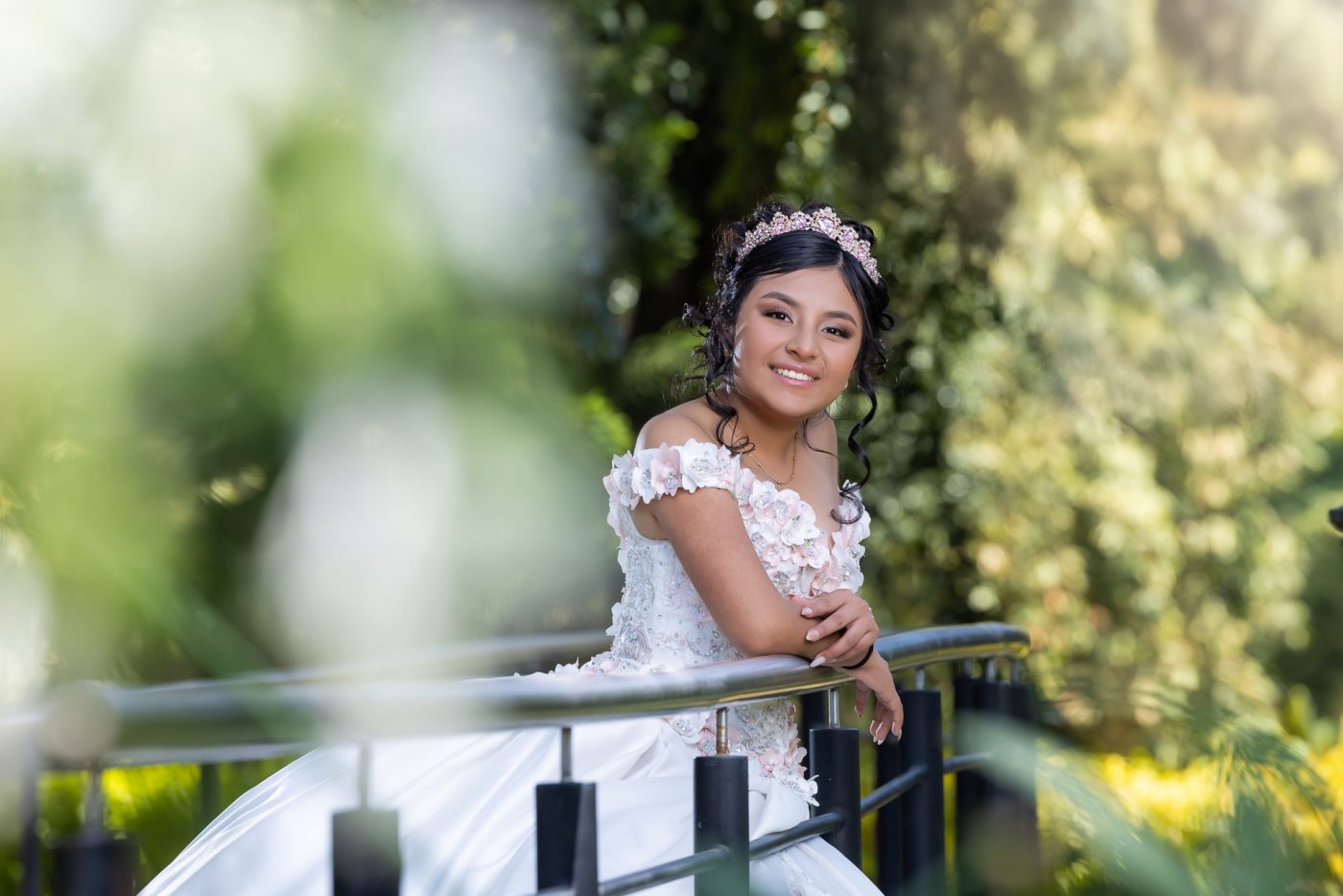 Retrato cercano de quinceañera sonriendo, recargada en un barandal con fondo verde desenfocado en los jardines de Los Pinos.