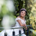 Retrato cercano de quinceañera sonriendo, recargada en un barandal con fondo verde desenfocado en los jardines de Los Pinos.
