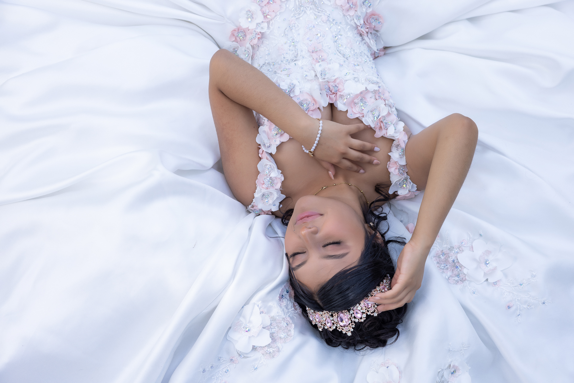 Toma cenital creativa de quinceañera acostada sobre la amplia falda de su vestido blanco con flores rosas, mirando hacia el cielo.
