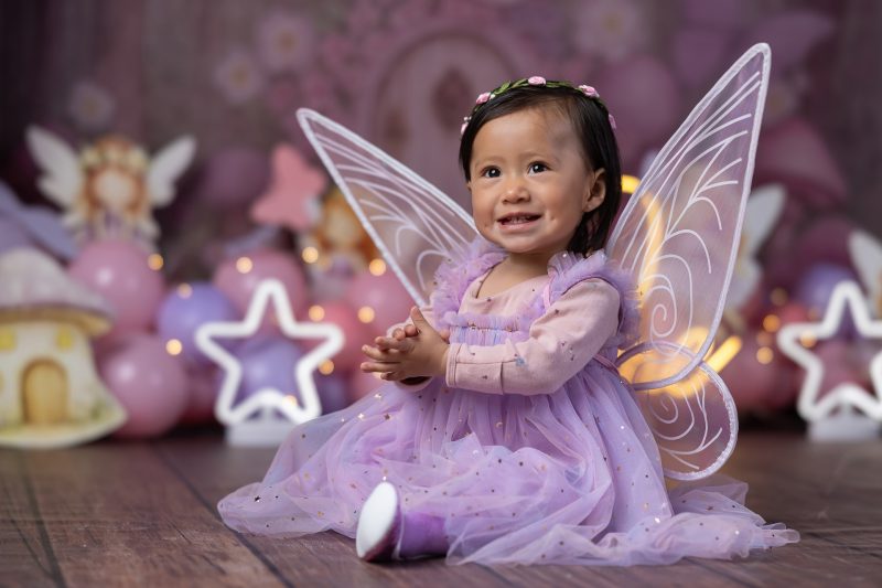 Primer plano de bebé sonriendo con alas de hada y vestido morado en decoración de bosque encantado