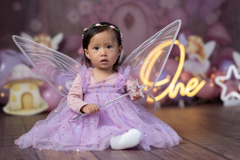 Bebé sentada sosteniendo una varita mágica con diadema de flores y alas de hada en estudio fotográfico