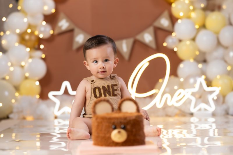 Niño sentado frente a pastel con forma de oso usando un traje de pana que dice ONE con decoración de luces y globos