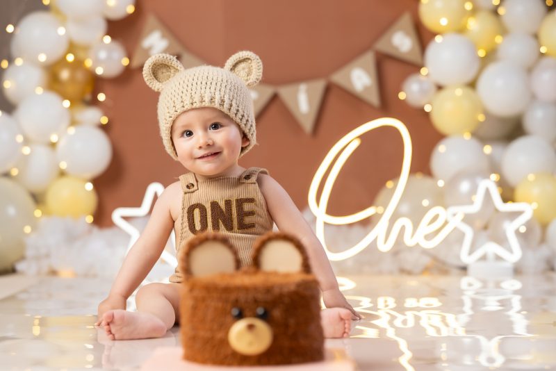 Bebé sonriente usando un gorro tejido de osito y overol beige frente a un pastel de oso en sesión Smash Cake