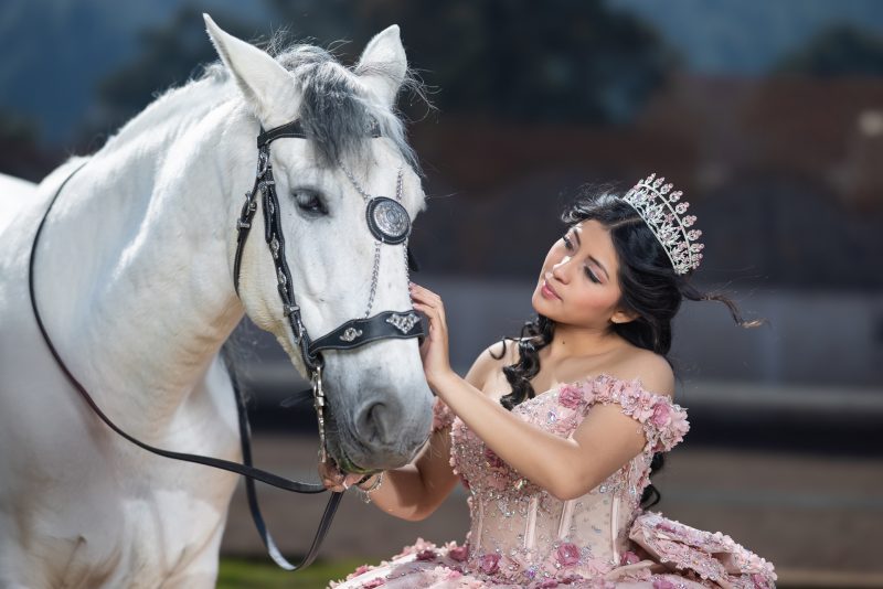 Primer plano de una quinceañera con tiara y un elegante vestido rosa con aplicaciones florales, acariciando suavemente la cabeza de un caballo blanco con brida decorada