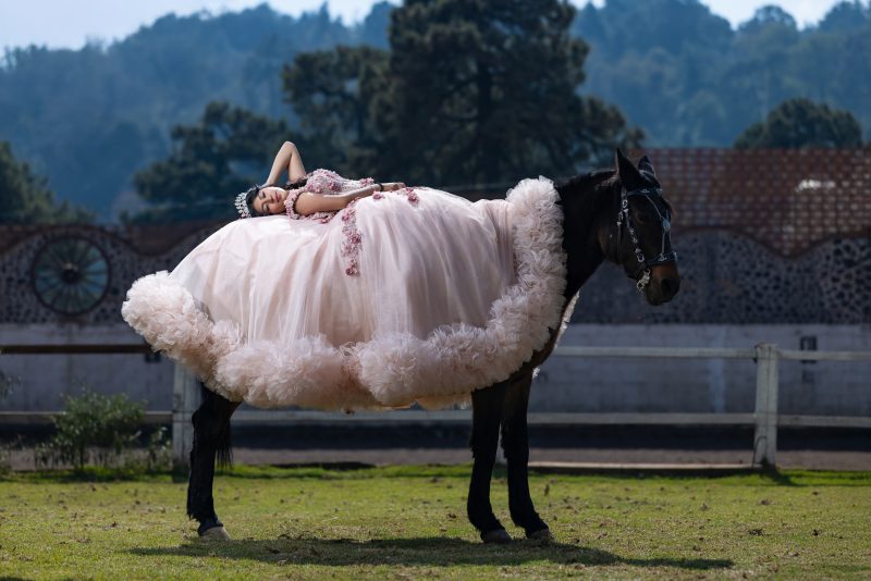 Pose artística y dramática de una quinceañera con su gran vestido rosa acostada sobre el lomo de un caballo oscuro, con el paisaje del rancho de fondo.