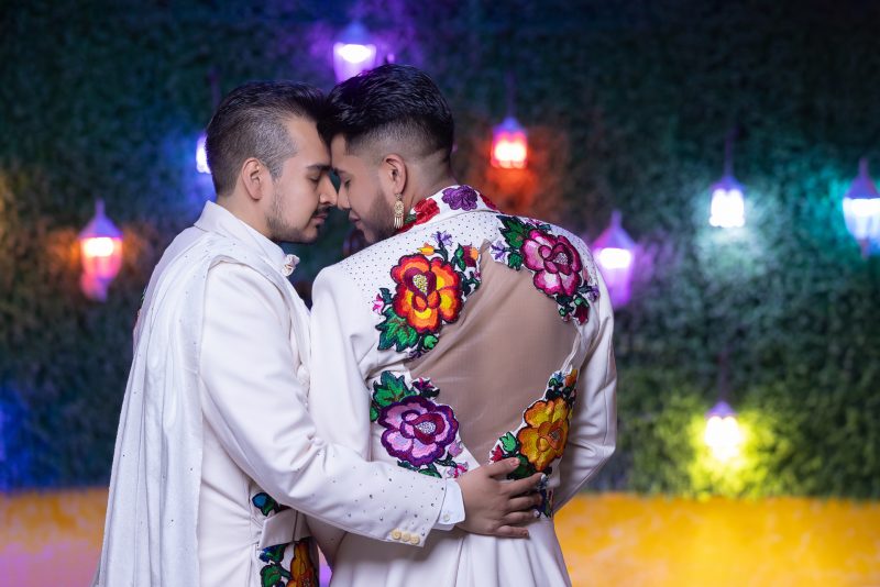 Primer plano emotivo de dos novios uniendo sus frentes con los ojos cerrados. Detalle de la espalda del saco con tela transparente y bordado de flores mexicanas rojas y rosas.
