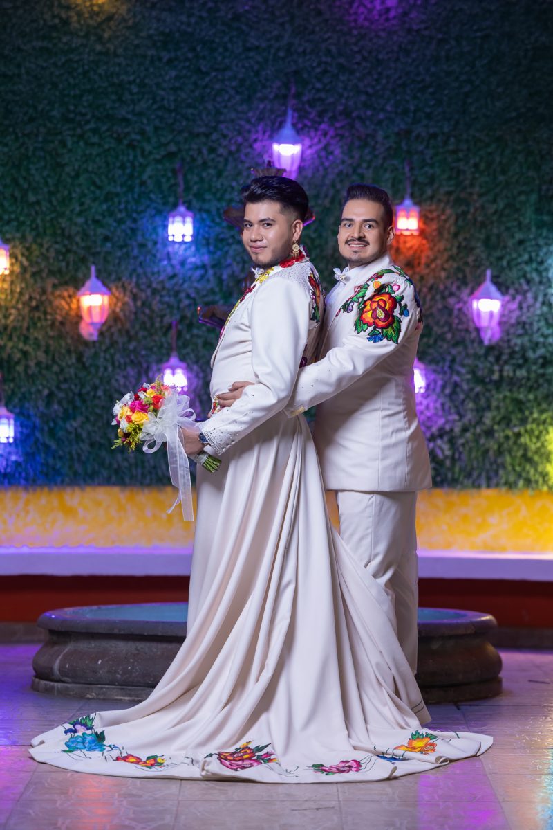 Pareja de novios gay posando de cuerpo completo espalda con espalda. Visten trajes blancos con bordados florales coloridos estilo mexicano; uno luce una falda/cola larga nupcial. Fondo de pared verde con luces decorativas.
