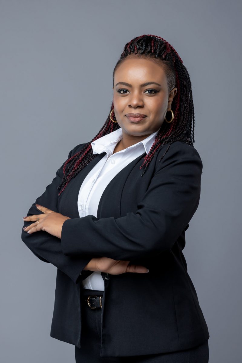 Retrato corporativo de mujer profesional con traje negro y peinado trenzado en fondo gris neutro.