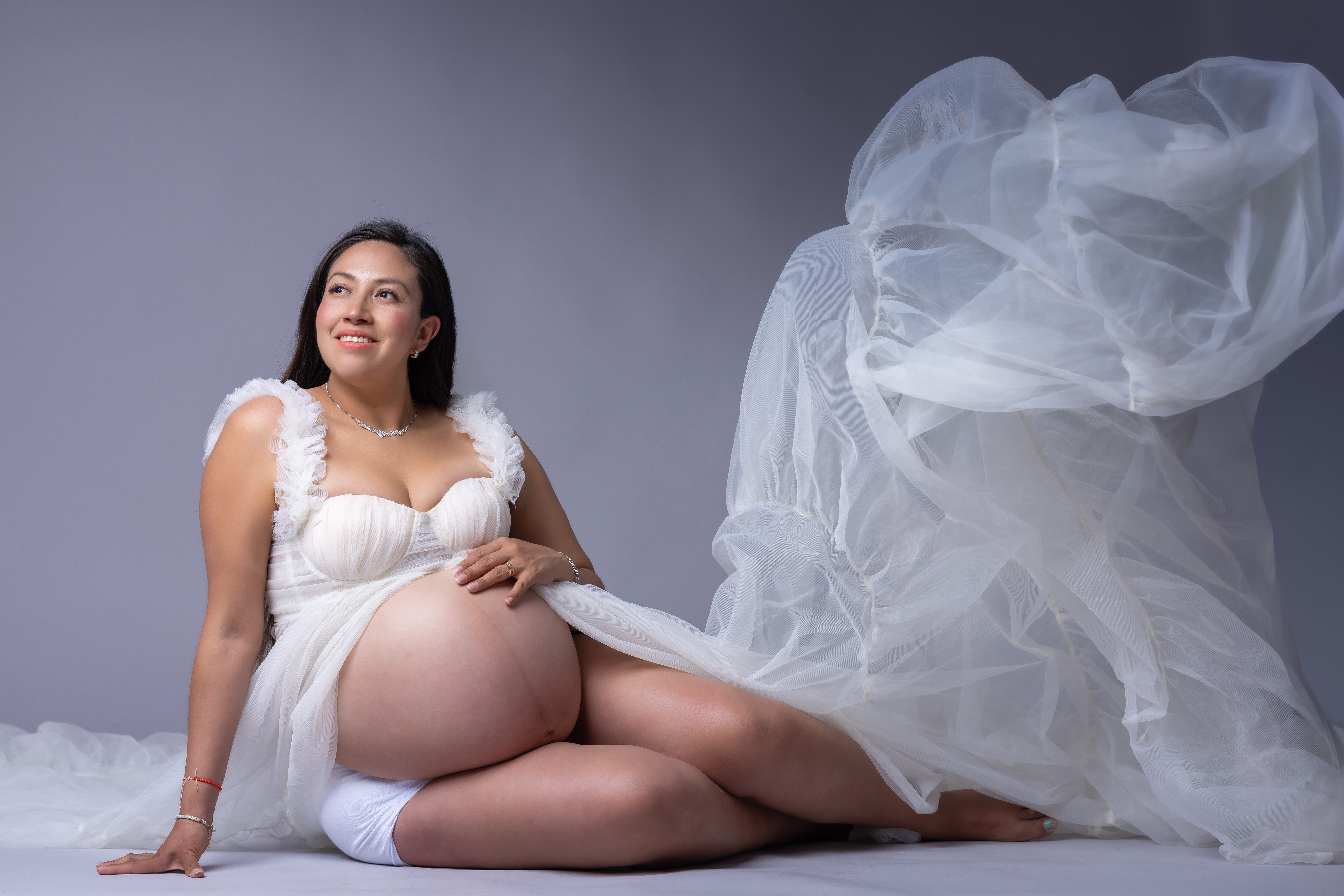 Mujer embarazada sonriendo sentada en el suelo de un estudio fotográfico, vistiendo un conjunto blanco con una tela de tul vaporosa que flota a su alrededor, mostrando su vientre descubierto sobre un fondo gris neutro.