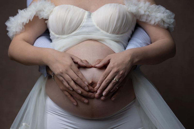 Primer plano del vientre de una mujer embarazada con las manos de ella y su pareja entrelazadas formando un corazón sobre la barriga, ella viste top blanco y él camisa azul a rayas.